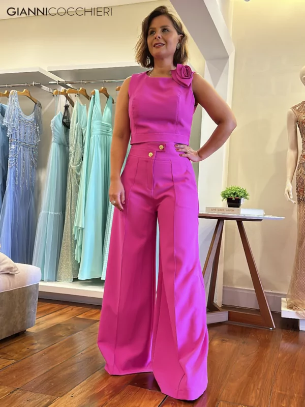 Conjunto de blusa e calça pantalona na cor fucsia. A camisa não possui mangas, contém uma flor no ombro esquerdo e tem o corte no estilo alfaiataria. A calça é cintura alta com dois botões e estilo pantalona.